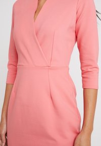 Femme portant une robe ajustée, rose clair, longueur genou, avec des manches trois-quarts et un corsage croisé en forme de col en V.