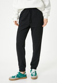 Zwarte sweatpants met een elastische tailleband en boorden, met een gladde textuur en schuine naaddetails. Gecombineerd met teal en grijze sneakers.