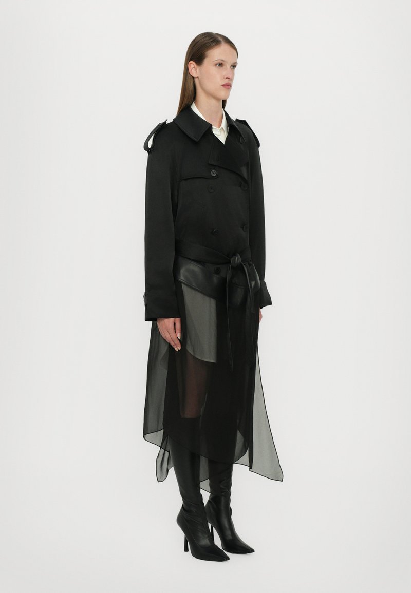 Trench coat nero con sovrapposizione trasparente, design doppiopetto, cintura in vita e stivali con tacco a punta. Tessuto dalla texture liscia.