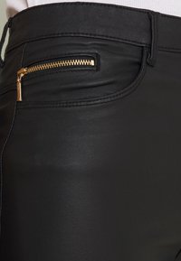 Gros plan d'un pantalon en cuir noir avec une petite poche zippée dorée et des coutures visibles près de la taille et des hanches.