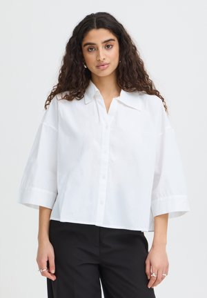 Młoda kobieta z długimi kręconymi włosami, ubrana w luźną białą koszulę na guziki i czarne spodnie, stojąca na jednolitym tle.