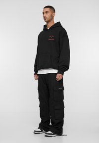 Black hoodie with front pocket and red text, paired with black cargo pants featuring multiple pockets and a patch, and black and white sneakers.