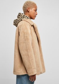 Bežový kabát z umelého kožúška s golierom, ktorý má kapucňu so leopardím vzorom. Dizajn je oversized, s jemnou textúrou a uvoľneným strihom.