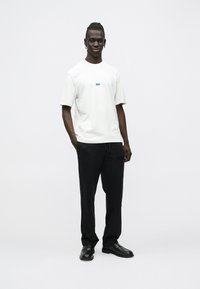 Άνδρας που στέκεται με τα χέρια στις τσέπες, φοράει λευκό κοντομάνικο πουκάμισο και μαύρο παντελόνι με μαύρα επίσημα παπούτσια, μπροστά σε απλό ανοιχτόχρωμο φόντο.