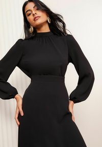 Černé šaty s dlouhými, nabíranými rukávy a vysokým výstřihem. Mají úzký pas a hladkou texturu, zdůrazněnou elegantní siluetou.
