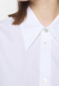 Vit skjortan med knappar framtill har en klassisk krage, plastknappar och en slät bomullstextur, och visar en minimalistisk design.