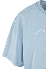 T-shirt en coton bleu clair avec un col rond, des manches courtes et un texte blanc brodé sur la poitrine. Texture lisse avec une coupe décontractée.