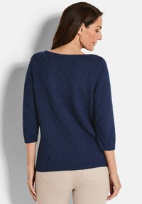 Dunkelblauem gestrickter Pullover mit Dreiviertelärmeln, der ein strukturiertes Muster und einen gerippten Saum aufweist. Trägt eine helle Hose.