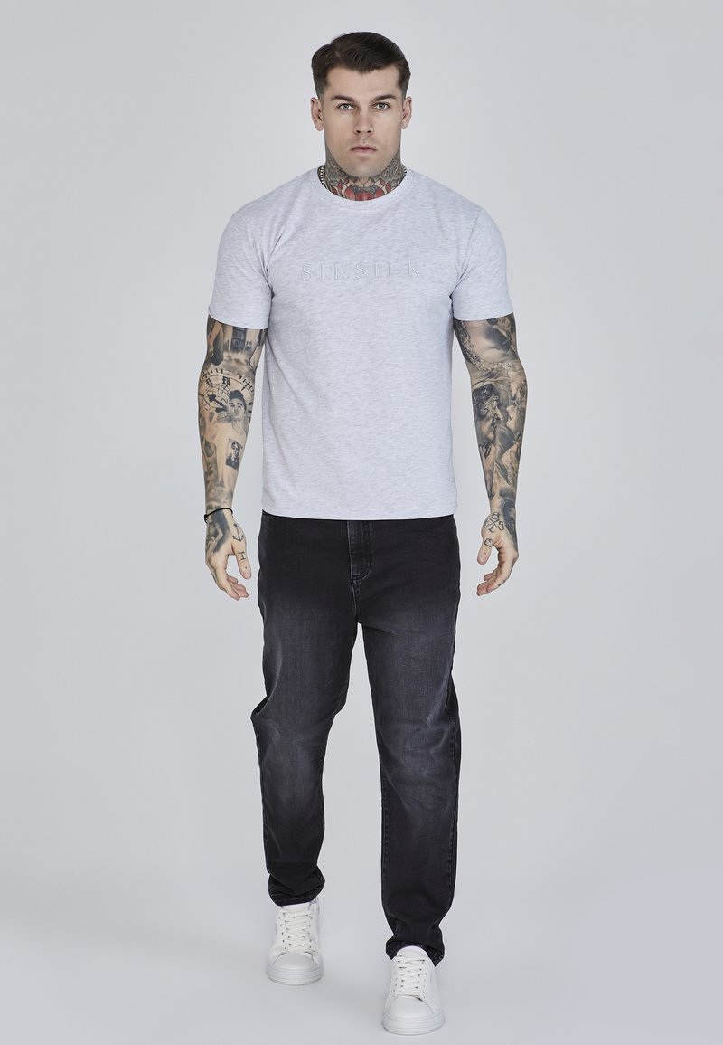 Maglietta grigia a maniche corte con logo in rilievo, abbinata a jeans neri e sneakers bianche. Tatuaggi visibili sulle braccia. Sfondo semplice.