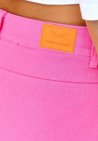 Różowa dżinsowa tkanina o teksturowanym wykończeniu, z kwadratową pomarańczową metką zawierającą tekst "stwórz siebie" wszytą w pas.