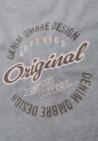 Tela azul claro con un diseño gráfico vintage que presenta las palabras "Original", "Diseño Ombre de Denim" y "Vintage City Co." en varias tipografías.