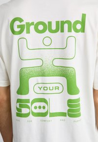 Hvid t-shirt med stort grønt grafisk tekst: "Ground YOUR SOLE" og abstrakte figurer, med "Nike for komfort og støtte" nedenunder.
