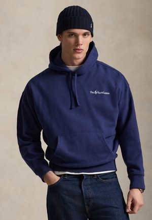 Giovane uomo che indossa un berretto di lana blu navy, una felpa con cappuccio Polo Ralph Lauren blu navy con logo, una maglietta bianca sotto e jeans blu scuro, posando su uno sfondo neutro.
