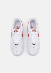 Fehér bőrből készült Nike Air Force 1 tornacipő piros részletekkel, perforált orr résszel, lapos fűzőkkel, valamint a nyelvén és a sarkán domború logóval.