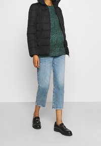 Chaqueta de plumas negra, parte superior verde con patrón, jeans cortos de mezclilla azul claro y mocasines negros con detalle de hebilla. Fondo neutro.