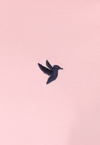 La tela de punto rosa claro presenta un pequeño logotipo de pájaro bordado en marino en el lado izquierdo. La textura es suave y ligeramente elástica.
