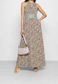 Robe longue à motif floral vert, avec un col halter et une taille froncée. Associée à un sac à main violet clair et des chaussures blanches.