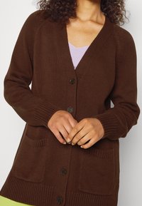 Hnědý pletený kardigan s výstřihem do V, zapínáním na knoflíky a dvěma předními kapsami. Manžety a spodní lem s žebrovaným vzorem. Lehká a texturovaná látka.