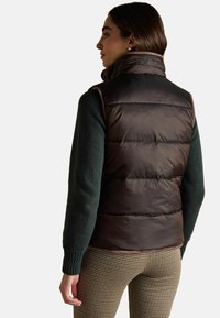 Tmavě hnědá péřová vesta s vysokým límcem, s prošívaným designem a detailem loga. Kombinována se zeleným svetrem a kalhotami s houndstooth vzorem.