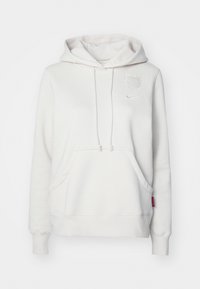 Haut à capuche blanc avec une poche avant, une capuche à cordon, un logo en relief et une étiquette rose. Texture douce, coupe décontractée et poignets côtelés.