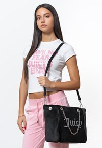 Zwart suede handtas met een zilveren kettinghandvat, voorzien van het woord "Juice" in rhinestones. De tas heeft een gestructureerde vorm en een platte basis.