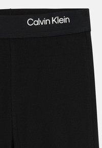 Les leggings noirs à côtes présentent une taille élastique lisse ornée du texte "Calvin Klein" en blanc. Le matériau semble doux avec une finition texturée.