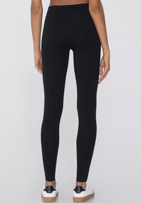 Leggings noirs en tissu extensible, présentant une taille haute et une texture lisse. Portés avec des baskets blanches et noires.