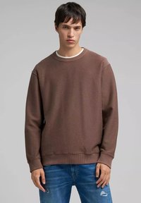 Sudadera marrón con acabado texturizado, cuello redondo y puños acanalados, combinada con jeans azules desgastados. Diseño sencillo y casual.