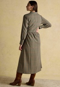 Garšs skotu raksta kleita zaļos un brūnos toņos, ar izdilušu jostu, garām piedurknēm un pogātu apkakli. Valkāta ar brūnām papēžu zābakiem.