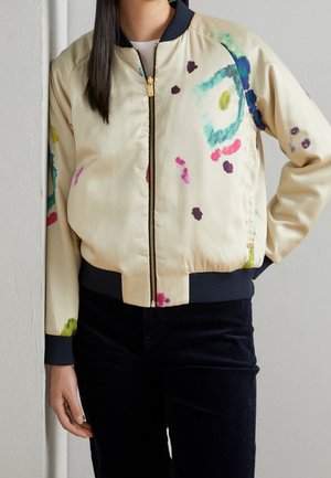 Personne portant une veste bomber beige zippée avec des poignets noirs et des motifs abstraits éclaboussés de peinture colorée, associée à un pantalon noir.