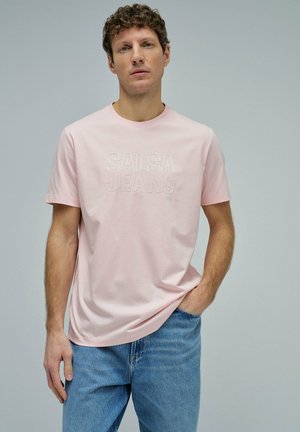 Hombre de pie con una camiseta rosa claro con el texto "SALSA JEANS" y vaqueros azules, mano en el bolsillo, fondo gris neutro.
