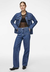 Giacca di jeans con due tasche anteriori, abbinata a jeans blu a vita alta. La texture è liscia, il colore è blu scuro. Footwear è nero.