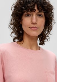 Lichtroze T-shirt van zachte stof, met een ronde halslijn en een kleine zak aan de linkerkant. Krullend haar deels zichtbaar.