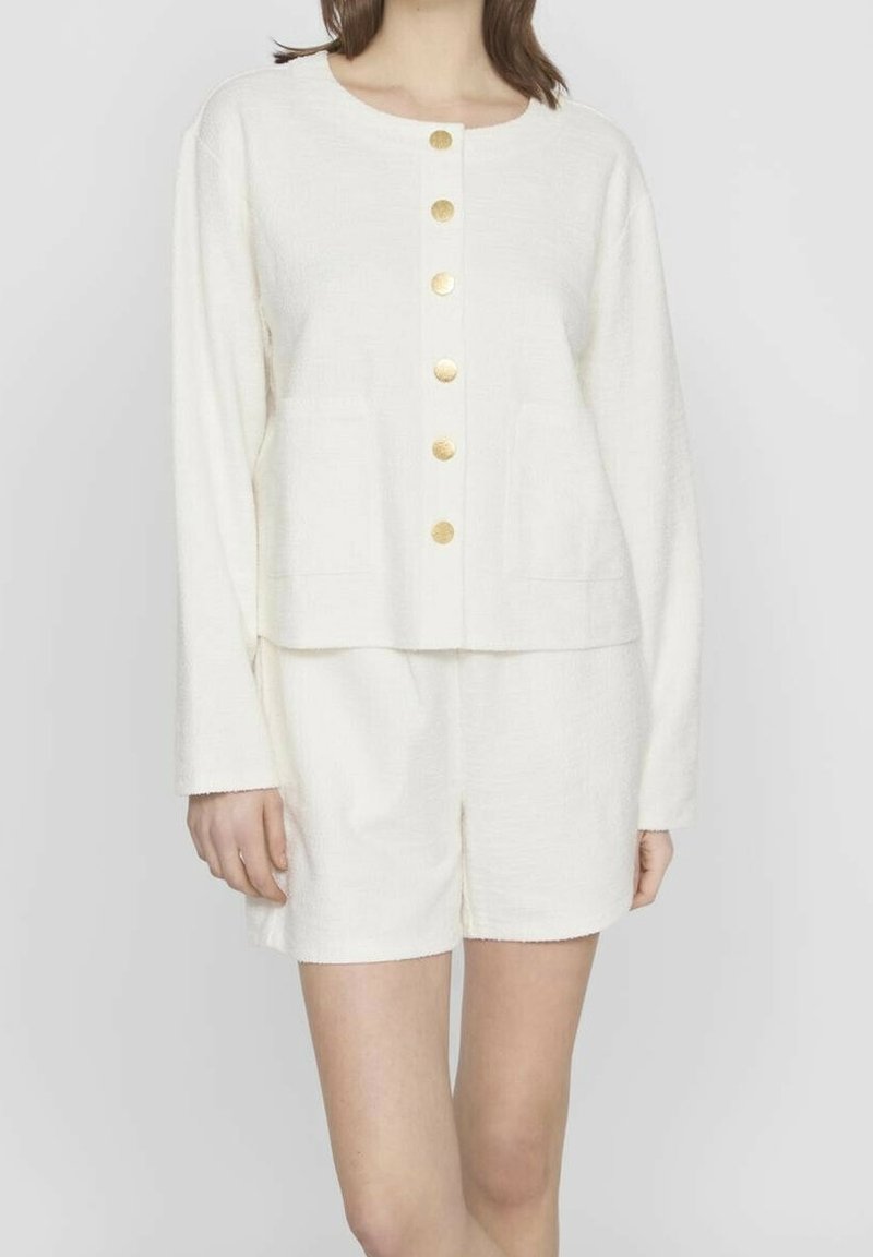Femme portant un cardigan blanc à manches longues boutonné avec des boutons dorés et un short blanc assorti, debout devant un fond uni.