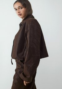 Brązowa skórzana kurtka o skróconym kroju, z zamkiem z przodu i kołnierzem. Gładka tekstura z lekko błyszczącym wykończeniem. Minimalistyczny styl.