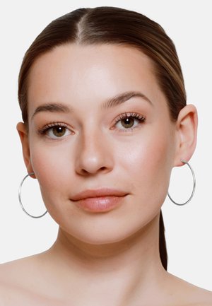 Młoda kobieta z gładkimi brązowymi włosami zaczesanymi do tyłu, nosząca duże srebrne kolczyki koła, neutralny makijaż i spokojny wyraz twarzy na białym tle.