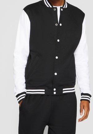 Mann trägt eine schwarz-weiße Collegejacke mit weißen Ärmeln und schwarzen Hosen, Hände teilweise in den Taschen vor hellem grauem Hintergrund.