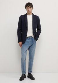 Mann, der gegen eine weiße Wand steht, trägt einen marineblauen Blazer, einen weißen Pullover, hellblaue Jeans und schwarze Slipper, mit einer Hand in der Tasche.