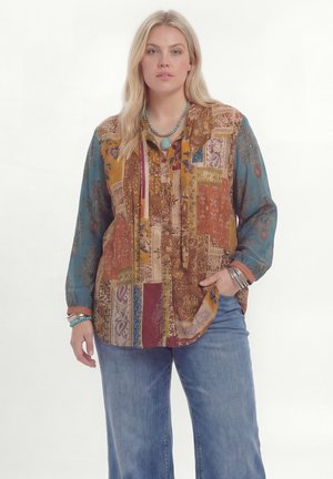 Femme portant un chemisier patchwork multicolore avec des manches bleues, un jean bleu, un collier turquoise et des bracelets argentés, debout devant un fond blanc.