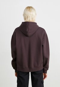 Mørkebrun oversized hettegenser med kengurulomme og snøring i hetten. Mykt bomullsstoff med ribbestrikkede mansjetter og nederkant.