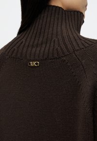 Maglione marrone lavorato a maglia con colletto alto a coste. Presenta un accento con logo dorato sul retro, che mette in mostra un tessuto texturizzato e una vestibilità rilassata.