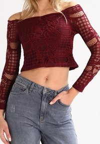 Žena v burgundské krajkové crop topu s odkrytémi rameny a vysokými modrými džíny, s jednou rukou v kapse, na jednobarevném pozadí.