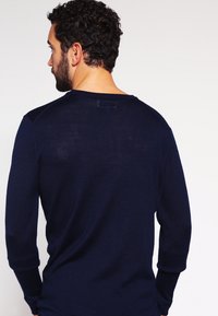 Marineblå stickad tröja med långa ärmar, ribbade muddarna och en vid halsringning; har en texturerad yta och subtil märkning på baksidan.
