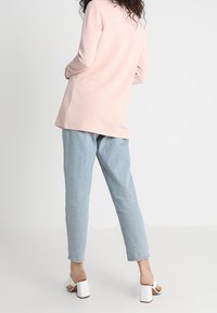 Ljusrosa långärmad topp i ribbad textur, tillsammans med ljusblå tapered jeans och vita blockklacksandaler, sedd från baksidan.