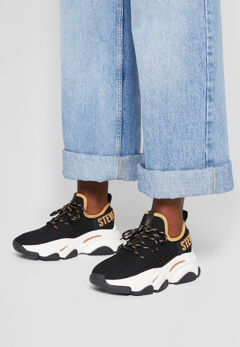 Baskets noires avec une tige en mesh texturé, accents orange et semelles blanches épaisses. Comprend une languette de talon avec logo et des lacets à motifs.