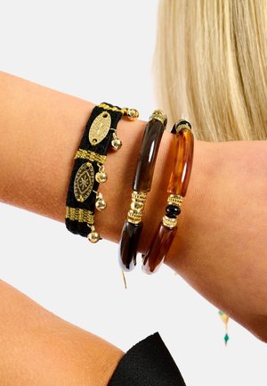 Trois bracelets : un bracelet noir tissé avec des accents dorés et des clochettes, ainsi que deux bracelets rigides en résine brillante aux tons marron et ambre avec des détails dorés.