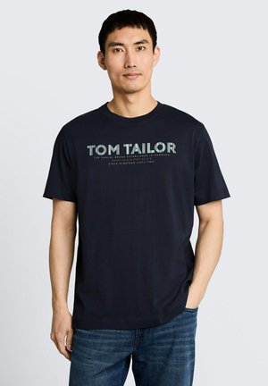 Man staat en draagt een zwart Tom Tailor t-shirt met logo tekst en blauwe jeans, een hand in de zak tegen een effen lichte achtergrond.