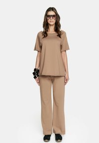 Casualny strój składający się z luźnej beżowej bluzki i dopasowanych szerokich spodni, uzupełniony czarnymi sneakersami i masywnymi czarnymi bransoletkami.