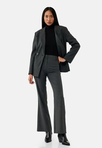 Grijze op maat gemaakte suit met een dubbelknoopblazer, een hoogsluitend zwart ribbelshirt en flairbroeken, gecombineerd met zwarte enkellaarsjes.