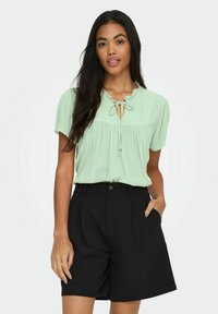 Hellgrüne Bluse mit kurzen Ärmeln, Krawattenkragen und Raffung, kombiniert mit hochgeschnittenen schwarzen Shorts mit Seitentaschen.